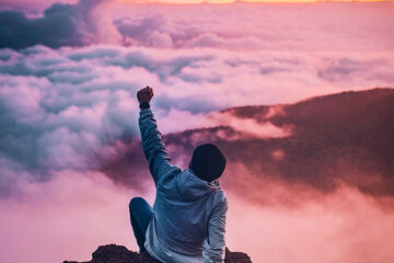 Person som sitter på ett berg över molnen med näven i luften för att fira sitt åstadkommande. SteadyBlogg