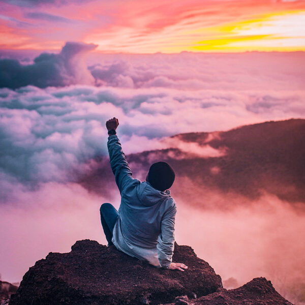 Person som sitter på ett berg över molnen med näven i luften för att fira sitt åstadkommande. SteadyBlogg